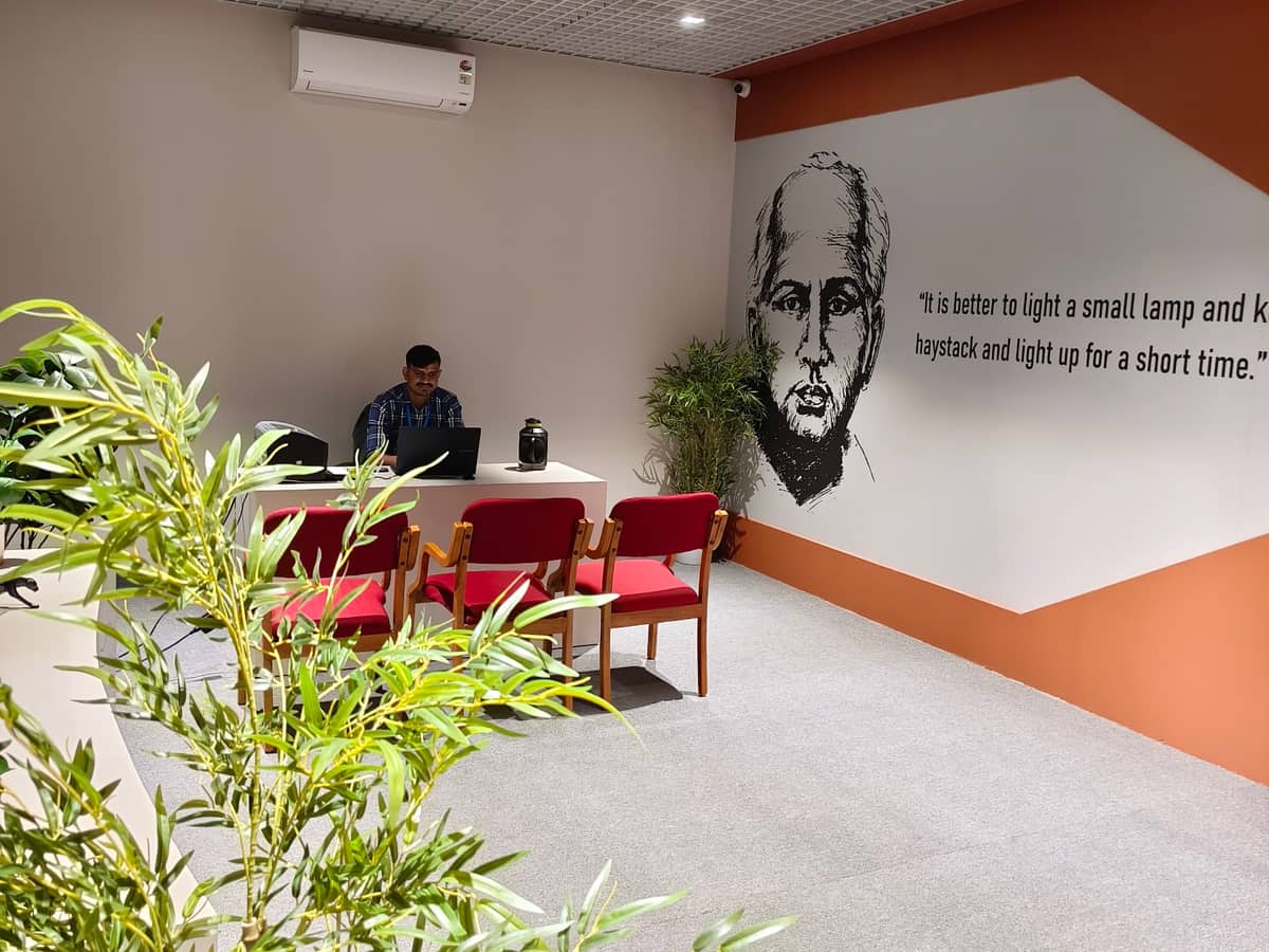 The front desk, with three red chairs in front of it, a person working at a laptop behind it, and a wall mural with the words: It is better to light a small lamp