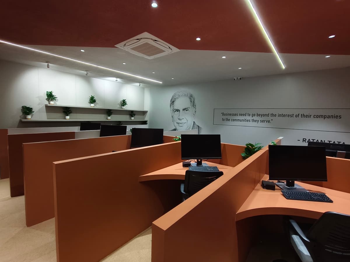Rows of orange workstation desks with monitors and keyboards, beneath a wall mural of Ratan Tata and his words on businesses serving their communities